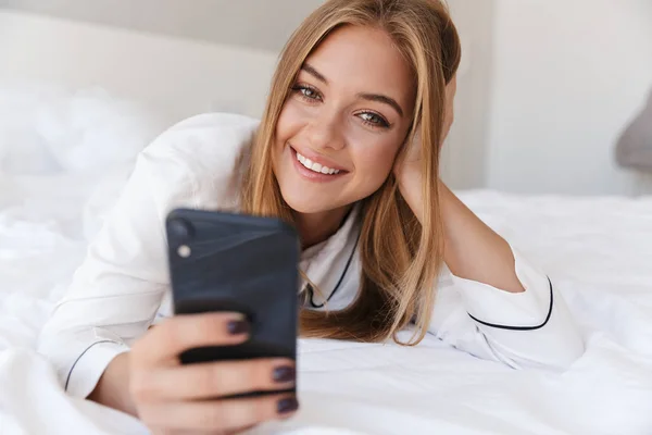 Photo of cheerful woman using cellphone and smiling while lying on bed