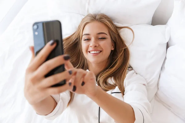 Foto de la encantadora mujer alegre usando el teléfono celular y sonriendo mientras miente —  Fotos de Stock