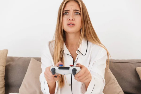 Foto de una mujer rubia infeliz jugando videojuegos mientras está sentada en el sofá —  Fotos de Stock