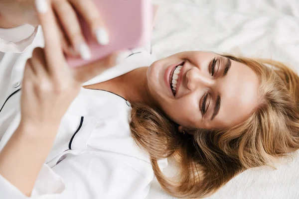Imagen de la joven feliz sosteniendo el teléfono celular mientras está acostado en la cama un —  Fotos de Stock