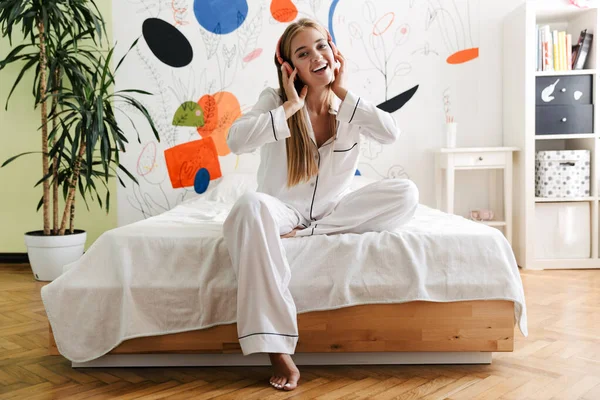 Image of caucasian young girl in headphones sitting on bed in ap — ストック写真