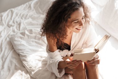 Yatakta iç çamaşırı giyen ve kitap okuyan bir kadın resmi.