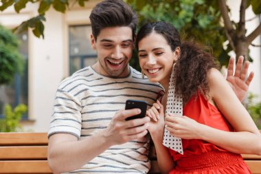 Heyecanlı genç bir çiftin otururken cep telefonu kullanıp kucaklaşması.