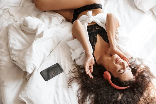 Image of woman wearing headphones lying on bed with smartphone a — ストック写真