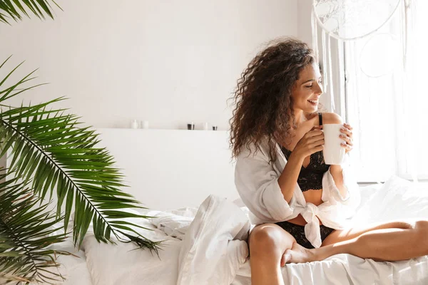 Immagine di una giovane donna rilassata che beve tè sul letto in un appartamento bianco — Foto Stock