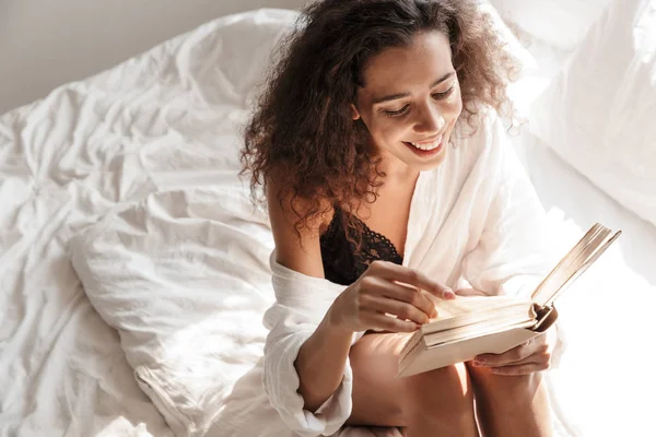 Image de la femme souriante et le livre de lecture tout en étant assis sur le lit dans — Photo