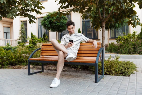 Imagen de un joven feliz usando un teléfono inteligente y sonriendo mientras está sentado — Foto de Stock