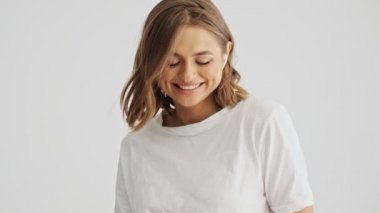 Beyaz tişörtlü, rahat gülümseyen genç bir kadın beyaz arka planda izole edilmiş kablosuz kulaklıklarıyla müzik dinliyor.