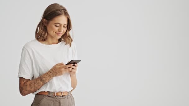 白いシャツを着ている楽観的な若い女性は 白い背景の上側に隔離された側面を見ながら 彼女のスマートフォンを使用して — ストック動画
