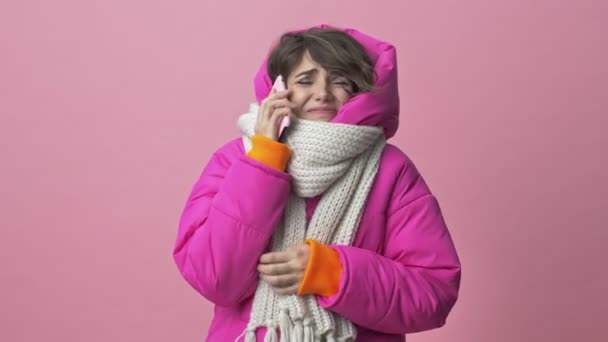スカーフで冬のジャケットを着た冷凍の若い女性がピンクの背景に隔離されたスマートフォンを呼び出しています — ストック動画
