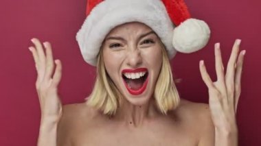 Kırmızı rujlu kızgın genç bir kadın Noel Baba şapkası takıyor. Çığlık atıyor. Kırmızı arka planda ellerini kaldırıyor.