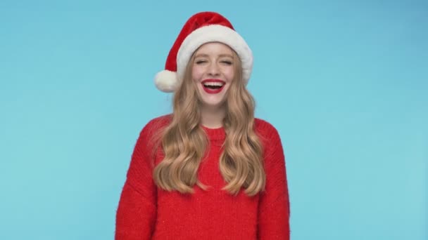 戴着圣诞礼帽的快乐而漂亮的女人笑着看着蓝色背景下的相机 — 图库视频影像