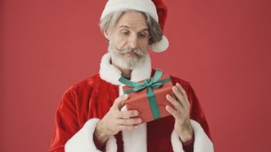 Noel Baba kostümlü neşeli, yaşlı, gri sakallı adam stüdyodaki kırmızı arka planda ne olduğunu tahmin ediyor.