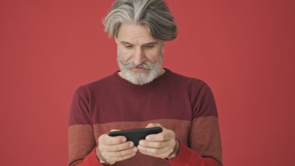 赤い壁に隔離されたゲームをプレイしながら スマートフォンを水平方向に保持赤いセーターの高齢者の白髪の髭の男を集中 — ストック動画