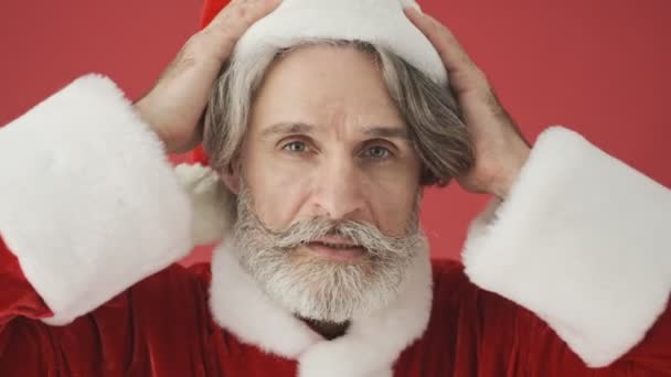身穿圣诞老人服装的英俊的白发苍苍的男人修正了他的红帽子 然后微笑着在工作室的红色背景下与摄像机隔离 — 图库视频影像