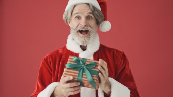 身穿圣诞老人服装的快乐的白发苍苍的老人送给摄像机一份礼物 摄像机被隔离在工作室的红色背景之外 — 图库视频影像