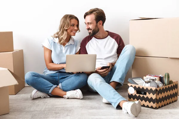 Foto de pareja alegre usando el ordenador portátil y el teléfono celular mientras está sentado —  Fotos de Stock