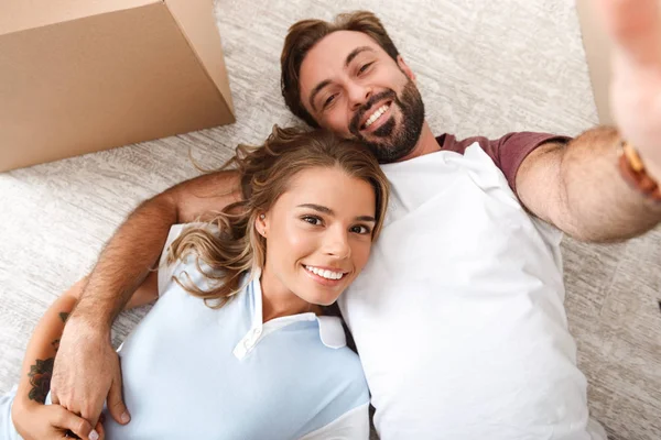 Photo of happy couple taking selfie photo and lying near cardboard box — 스톡 사진