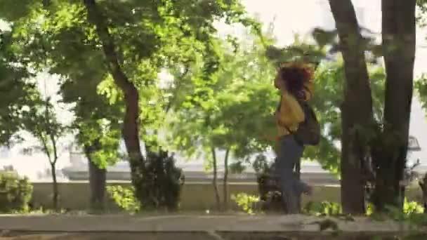 一个漂亮的年轻女人 一头卷曲的红头发 穿着黄色西装 牛仔裤 背包在绿地公园里奔跑 — 图库视频影像