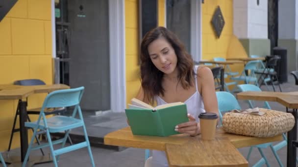 一个光彩夺目的年轻女子 带着稻草袋和一杯咖啡 坐在舒适的户外咖啡馆里看书 — 图库视频影像