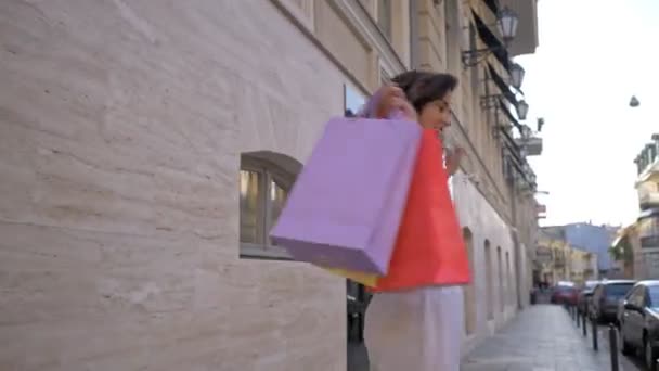 快乐微笑的年轻女子一边走在街上一边挥动着彩色购物袋 — 图库视频影像