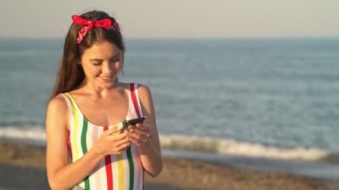 Kırmızı saç bandı ve çizgili mayo takan güzel bir kadın deniz kenarında dururken akıllı telefonuyla sohbet ediyor.