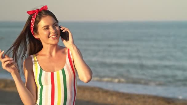 一位快乐的年轻女子 头戴红色头巾 身穿条纹泳衣 站在海边 一边打电话一边聊天 — 图库视频影像