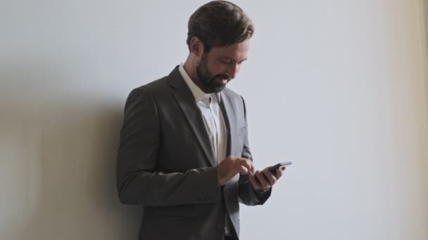 オフィスの壁の近くに立っている間 スマートフォンを使用してハンサムな髭のビジネスマンを喜ばせます — ストック動画