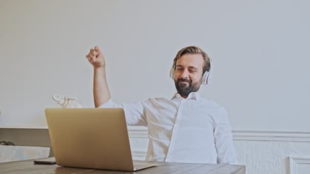 オフィスのテーブルに座っている間 楽しいとラップトップコンピュータを使用してヘッドフォンで陽気なハンサムなひげを生やしたビジネスマン — ストック動画