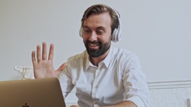 オフィスのテーブルに座っている間にノートパソコンでビデオ通話で話すハンサムな髭のビジネスマンを喜ばせます — ストック動画