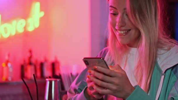 カラフルなネオンの壁の上にスマートフォンや飲み物カクテルを使用して楽しい若い美しい女性のビューを閉じます — ストック動画