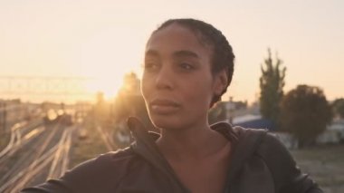 Odaklanmış Afro-Amerikan kadın spor giyim kuşamında eski köprü üzerinde çalışırken korkuluklara yaslanıyor.