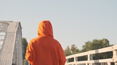 Kafasında turuncu kapüşonlu bir kapüşon olan atletik bir insanın arkadan görünüşü sabah köprüde koşuyor