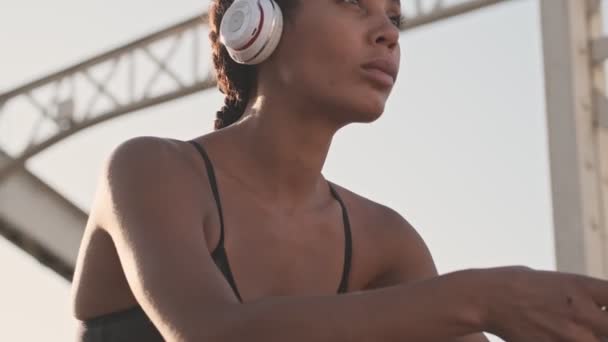 古い橋の上にワイヤレスヘッドフォンを持つスポーティーなアフリカ系アメリカ人女性の考え方をクローズアップ — ストック動画