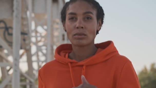 オレンジのパーカーを着たアフリカ系アメリカ人の女性が橋の上のカメラに向かって走っている — ストック動画