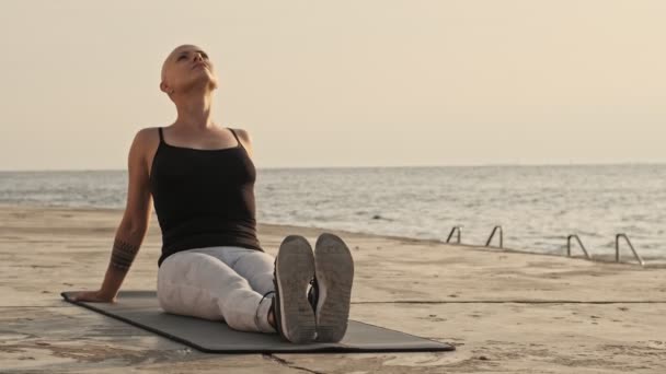海の近くのマットに座っている間に静かな魅力的なハゲスポーツの女性が休んで離れて見て — ストック動画
