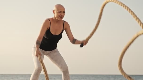 魅力的なはげスポーツ女性が海の近くにクロスフィットロープで運動を行う笑顔屋外 — ストック動画