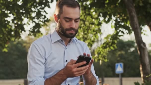 屋外の公園に座っている間にスマートフォンを使用して静かなひげそりのビジネスマン — ストック動画