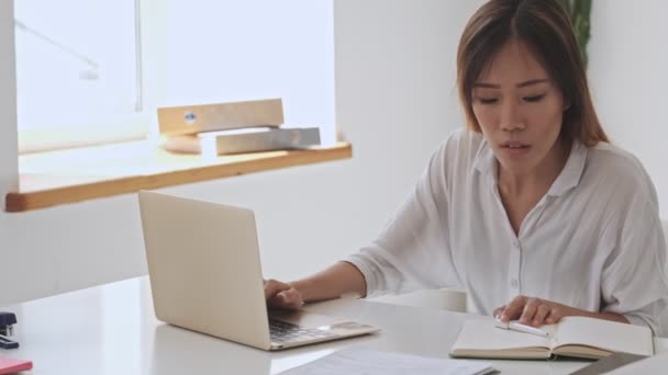 严肃的亚洲女商人坐在办公室的桌子边 一边拿着笔记本一边做笔记 — 图库视频影像