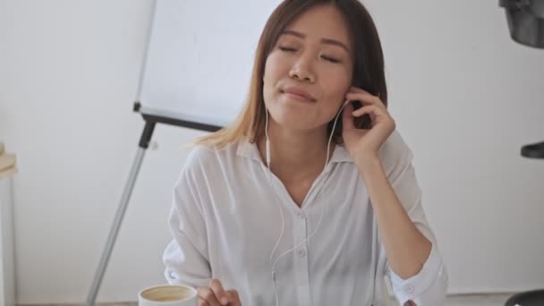 一位漂亮的亚洲女商人在白色的办公室里一边欣赏音乐 一边用她的有线耳机 — 图库视频影像