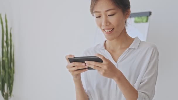 ホワイトオフィスでスマートフォンを水平方向に使いながら 幸せなアジアの女性のメッセージを笑顔 — ストック動画