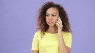 Sarı tişörtlü güzel, mutlu bir Afrikalı kadın akıllı telefondan konuşuyor ve mor arka plandan kameraya bakıyor.