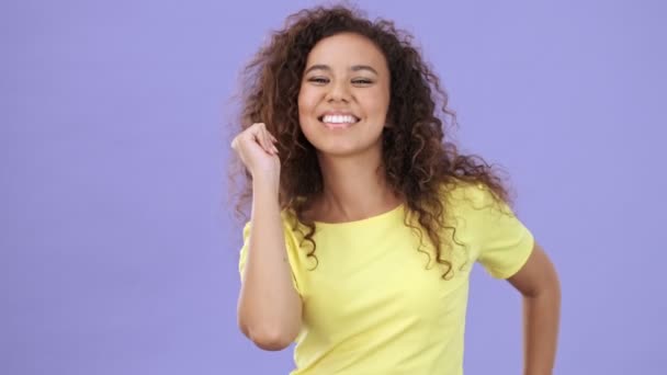 美しいです幸せなアフリカの若いです女性で黄色のTシャツダンスながら カメラを見ながら紫の背景孤立 — ストック動画