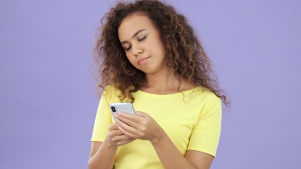 穿着黄色T恤的漂亮的非洲姑娘在与紫色背景隔离的智能手机上聊天时变得很不高兴 — 图库视频影像