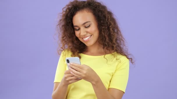 紫の背景の上にスマートフォンを使用している間 周りを見回す黄色のTシャツの思考幸せなアフリカの若い女性孤立 — ストック動画