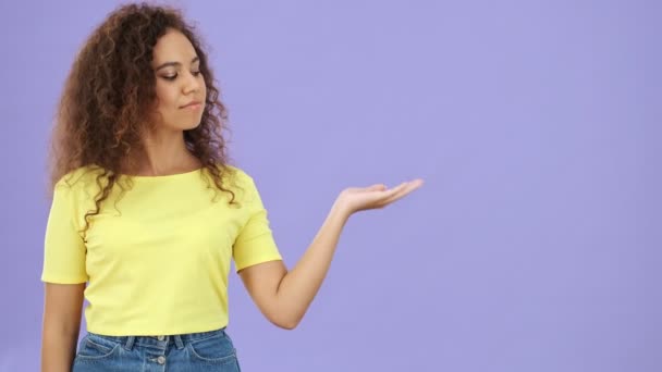 明るいアフリカの若いです女性で黄色のTシャツ指摘指でコピースペースで彼女の手のひら上にカメラを見ながら紫色の背景孤立 — ストック動画