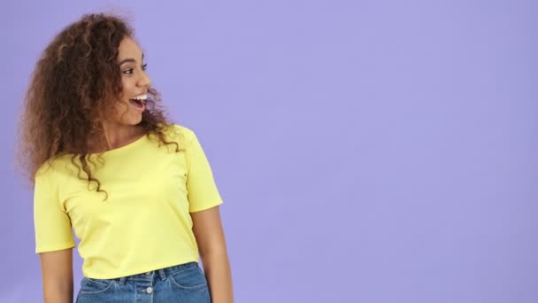 黄色のTシャツを着た魅力的なアフリカの若い女性は 紫色の背景に孤立した何かを比較しながら親指を横に向けてジェスチャー — ストック動画