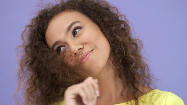 在镜头前 一个穿着黄色T恤的可爱的调情的非洲女青年微笑着玩着她的头发 在紫色的背景下凝视着相机 — 图库视频影像