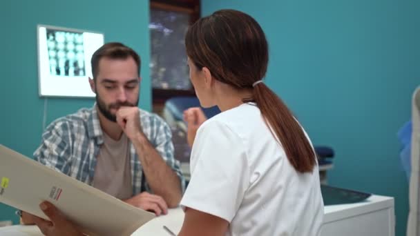 テーブルで病院で病歴を保持しながら集中治療の女医師が患者と話している — ストック動画