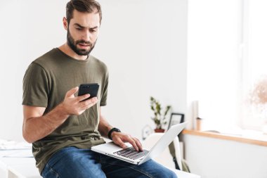 Cep telefonuyla yazı yazan ve dizüstü bilgisayar kullanan kafası karışmış yakışıklı bir adam resmi